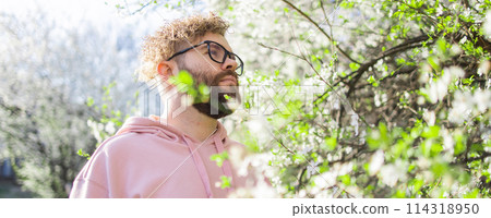 Banner Male bearded man standing under branches with flowers of blooming almond or cherry tree in spring garden. Spring blossom. Copy space 114318950