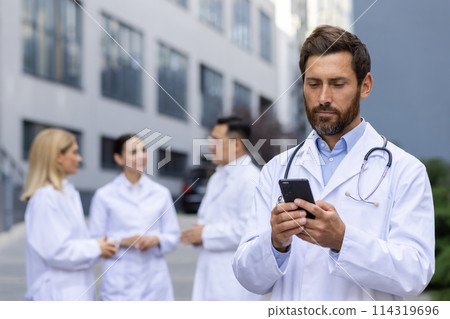 A serious young male doctor, scientist is standing outside a hospital room and is concentrating on using a mobile phone, colleagues are standing behind him and are looking at each other. 114319696