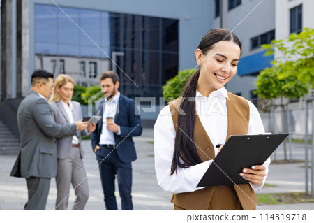 Businesswoman with four colleagues outside office building, company employee checking documents, smiling contentedly. Businesswoman with four colleagues outside office building, company employee checking documents, smiling contentedly. 114319718