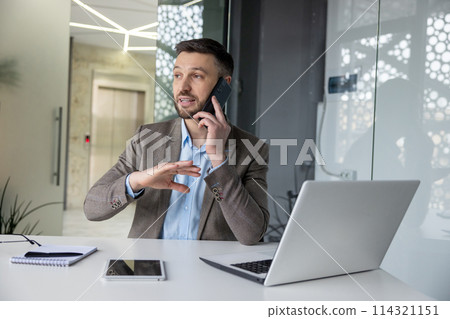 A professional businessman in a modern office setting communicates over the phone while gesturing actively, with a laptop and tablet in front of him. 114321151