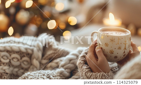 Person holding a coffee cup of hot chocolate in front of a Christmas tree Person holding a coffee cup of hot chocolate in front of a Christmas tree 114325079