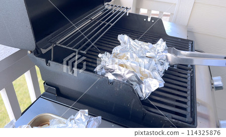 Several ears of corn wrapped in aluminum foil are lined up on a barbecue grill, slowly roasting to perfection, showcasing a popular and delicious method of cooking this classic side dish. 114325876