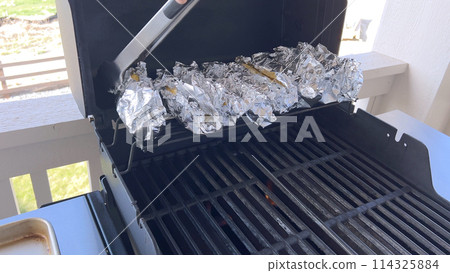 Several ears of corn wrapped in aluminum foil are lined up on a barbecue grill, slowly roasting to perfection, showcasing a popular and delicious method of cooking this classic side dish. 114325884