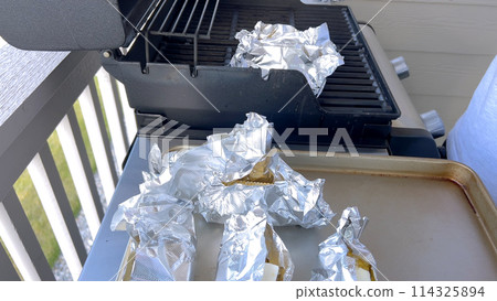 Several ears of corn wrapped in aluminum foil are lined up on a barbecue grill, slowly roasting to perfection, showcasing a popular and delicious method of cooking this classic side dish. 114325894