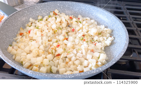 A frying pan on a gas stove sizzles with diced potatoes and colorful bits of red and green peppers, showcasing a delicious and simple home-cooked side dish in the making. 114325900