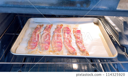 Crispy bacon strips bake evenly on a parchment-lined tray inside an oven, showcasing a clean and efficient method for achieving perfectly cooked bacon with minimal mess. 114325920