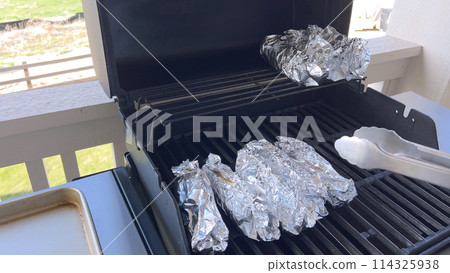 Several ears of corn wrapped in aluminum foil are lined up on a barbecue grill, slowly roasting to perfection, showcasing a popular and delicious method of cooking this classic side dish. 114325938