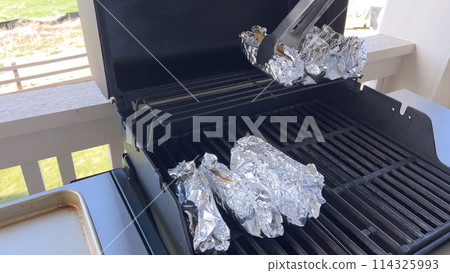Several ears of corn wrapped in aluminum foil are lined up on a barbecue grill, slowly roasting to perfection, showcasing a popular and delicious method of cooking this classic side dish. Several ears of corn wrapped in aluminum foil are lined up on a barbecue grill, slowly roasting to perfection, showcasing a popular and delicious method of cooking this classic side dish. 114325993