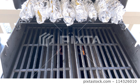 Several ears of corn wrapped in aluminum foil are lined up on a barbecue grill, slowly roasting to perfection, showcasing a popular and delicious method of cooking this classic side dish. 114326010