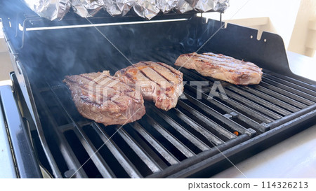 A person uses tongs to flip juicy steaks on a grill, with flames licking the meat, captured in a moment of intense heat and sizzle, showcasing a classic outdoor cooking experience. 114326213