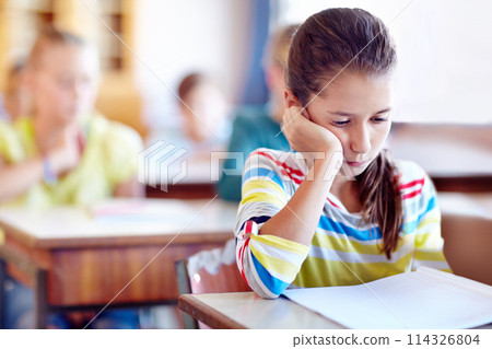 Child, school and desk with book or bored in classroom or education lesson or reading, knowledge or studying. Girl, kids and unhappy or learning or tired pupil with paper or fatigue, student or moody 114326804