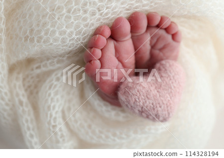 The tiny foot of a newborn baby. Soft feet of a new born in a white wool blanket. Close up of toes, heels and feet of a newborn. Knitted pink heart in the legs of a baby. Macro photography 114328194