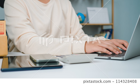 Woman runs an e-commerce business is checking orders from laptop, she owns an online store, she packs and ships through a private transport company. Online selling and online shopping concepts. 114328408