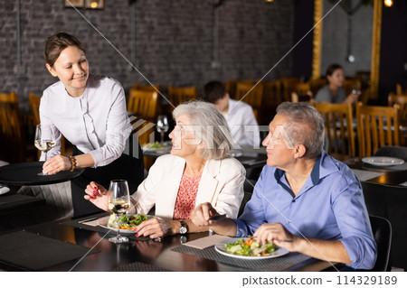 Waitress serving wine to senior couple having dinner in restaurant 114329189