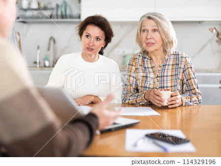Two women sitting at the kitchen-table face to face with male agent 114329196