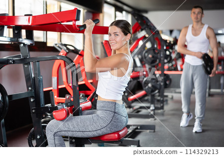 Girl doing strength training in gym, exercising upper body muscles on shoulder press machine 114329213