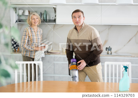Man vacuum-cleaning in the kitchen while his woman wiping plates kitchen 114329703