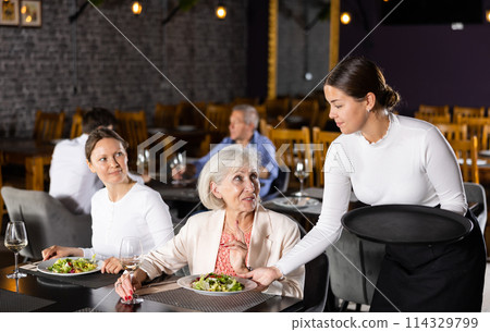 Smiling waitress fulfills order of young girl and elderly woman of restaurant visitors. Smiling waitress fulfills order of young girl and elderly woman of restaurant visitors. 114329799