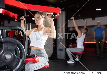 Girl doing strength training in gym, exercising upper body muscles on shoulder press machine 114329949