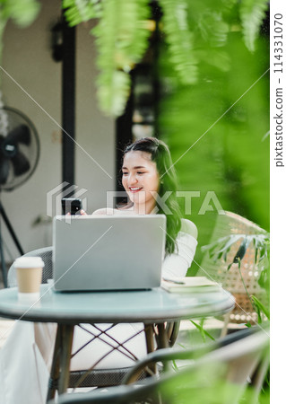 Smiling young woman enjoys checking her smartphone while seated with a laptop at a garden cafe, creating a harmonious work environment. 114331070