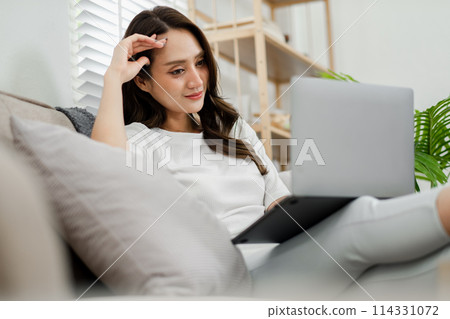 Woman reclines on a couch at home, working on a tablet with a thoughtful expression, surrounded by a peaceful indoor environment. Woman reclines on a couch at home, working on a tablet with a thoughtful expression, surrounded by a peaceful indoor environment. 114331072
