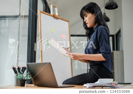 Concentrated young Asian businesswoman reviewing a financial report while standing at her office workspace. Concentrated young Asian businesswoman reviewing a financial report while standing at her office workspace. 114331099