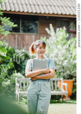 Smiling young woman enjoying remote work with her laptop in a green outdoor setting, embodying work flexibility. Smiling young woman enjoying remote work with her laptop in a green outdoor setting, embodying work flexibility. 114331100