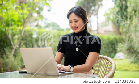 An attractive Asian woman sits at a table in a backyard using her laptop computer. 114331325
