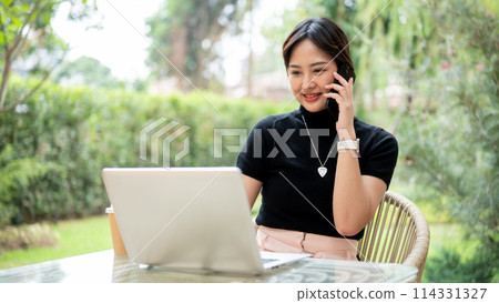 An attractive Asian woman sits at a table in a garden using her laptop and talking on the phone. 114331327