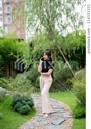 A positive, beautiful Asian woman talking on the phone while walking in a beautiful green garden. A positive, beautiful Asian woman talking on the phone while walking in a beautiful green garden. 114331331