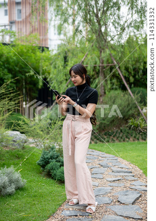 A positive Asian woman using her phone, chatting or checking messages while walking in a garden. 114331332