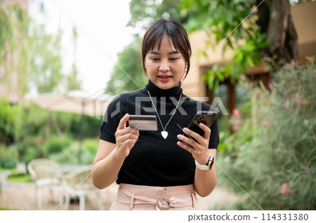 A beautiful Asian woman holding a credit card and a smartphone while walking in a beautiful garden. A beautiful Asian woman holding a credit card and a smartphone while walking in a beautiful garden. 114331380
