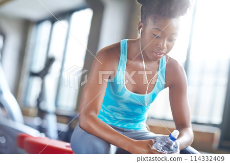 Fitness, earphone and black woman in gym, music and streaming audio in sportswear. Workout, sports and bottle for female athlete person, podcast or motivation in wellness facility for cardio exercise 114332409