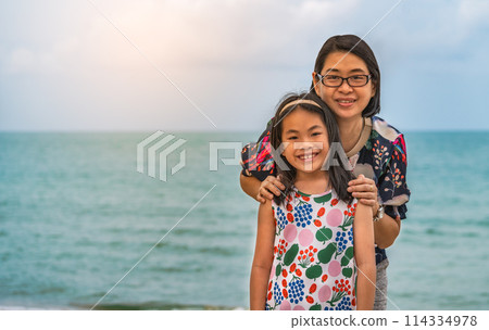 Portrait of a cute Asian child girl and a middle-aged mother on a beach, both are smiling and looking at the camera, blurred background of sea and sky, space for text,  landscape image. 114334978