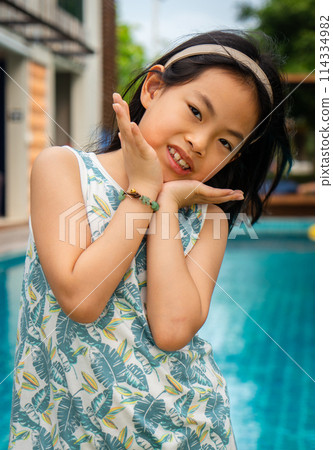 Portrait of an Asian cute girl with cute act in front of a resort swimming pool, girl in casual dress, eyes looking at camera, vertical image. 114334982