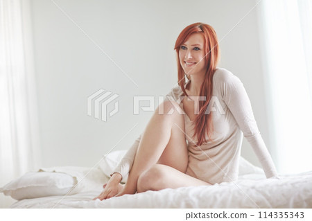 Woman, sitting and happy smile on bed for relax with morning, casual weekend and natural light in home. Redhead girl, rest and free time in bedroom in house with calm, wellness and peace in apartment 114335343