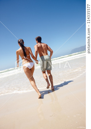 Couple, rear view and holding hands by beach with travel for bonding, summer vacation and anniversary getaway. Man, woman and love by ocean with swimwear for walking, healthy relationship or blue sky 114335577