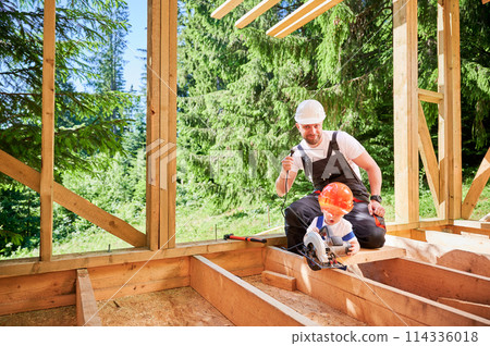Father with toddler son building wooden frame house. Man playing with his son using electric saw, demonstrating that it unplugged from power source on construction site. Carpentry and family concept. 114336018