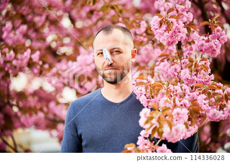Man allergic suffering from seasonal allergy at spring. Bearded man with clothespin clipped to his nose - symbolic gesture of his inability to breathe due to nasal congestion near blooming tree. Man allergic suffering from seasonal allergy at spring. Bearded man with clothespin clipped to his nose - symbolic gesture of his inability to breathe due to nasal congestion near blooming tree. 114336028