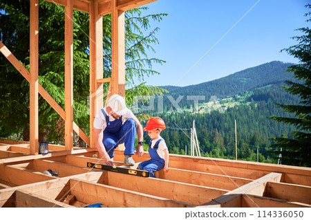 Father with toddler son building wooden frame house. Male builder demonstrating to his son how to measure level using spirit level on construction site on sunny day. Carpentry and family concept. 114336050