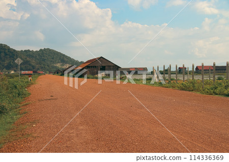A serene rural landscape with a dirt road leading past wooden salt storage houses and mountains in the background in Fish Island, Kampot Cambodia A serene rural landscape with a dirt road leading past wooden salt storage houses and mountains in the background in Fish Island, Kampot Cambodia 114336369