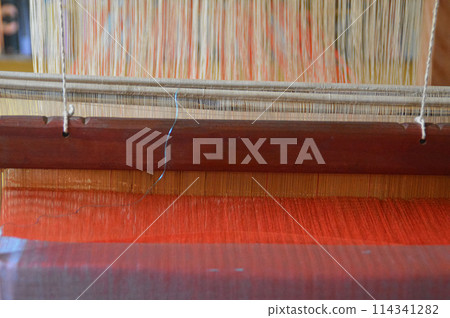 Close-up view of a traditional weaving loom with red and white cotton yarn threads that forms a weft of the fabric in the traditional technique of Laos called Ghot Close-up view of a traditional weaving loom with red and white cotton yarn threads that forms a weft of the fabric in the traditional technique of Laos called Ghot 114341282