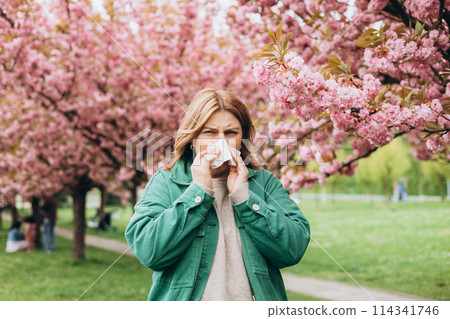 Sneezing young redhead woman with nose wiper among blooming trees in park. Portrait of sick women sneezes in white tissue, suffers from rhinitis and running nose. Symptoms of cold or allergy. Sneezing young redhead woman with nose wiper among blooming trees in park. Portrait of sick women sneezes in white tissue, suffers from rhinitis and running nose. Symptoms of cold or allergy. 114341746