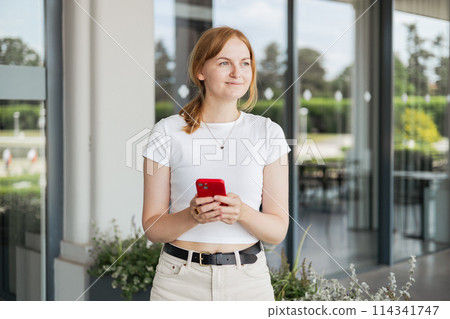 Smiling attractive young blonde woman wearing white t-shirt walking outdoors, using mobile phone. Urban lifestyle concept. Summer time 114341747