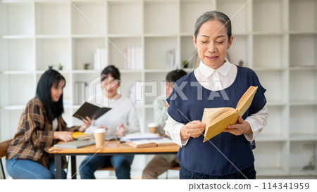 A smiling senior Asian female teacher professor standing in the classroom with a book in her hand. A smiling senior Asian female teacher professor standing in the classroom with a book in her hand. 114341959