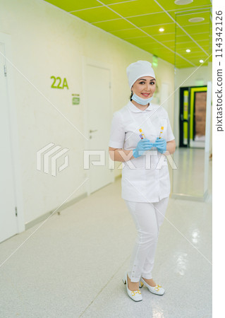 Full length vertical portrait of charming medical worker holding test tubes with blood standing on hallway in laboratory. Friendly nurse in medical uniform and blue gloves shows vials of liquid. 114342126