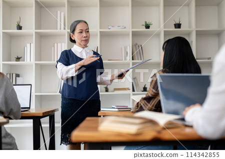 A professional mature Asian female teacher teaching students in the classroom, explaining a lesson. A professional mature Asian female teacher teaching students in the classroom, explaining a lesson. 114342855