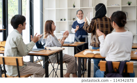 Positive mature Asian female teacher listening to her student presenting a project in the classroom. Positive mature Asian female teacher listening to her student presenting a project in the classroom. 114342878