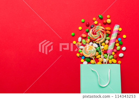 shopping paper gift bag in corner full of assorted traditional candies falling out on colored background with copy space. Happy Holidays sale concept shopping paper gift bag in corner full of assorted traditional candies falling out on colored background with copy space. Happy Holidays sale concept 114343253