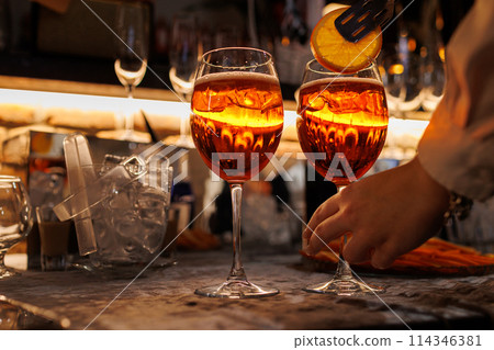 Bartender makes two glasses of cocktail Aperol spritz on bar counter, adds fresh orange slices. Typical alcoholic Italian beverage, aperitif made with Prosecco sparkling white wine and ice cubes. Bartender makes two glasses of cocktail Aperol spritz on bar counter, adds fresh orange slices. Typical alcoholic Italian beverage, aperitif made with Prosecco sparkling white wine and ice cubes. 114346381